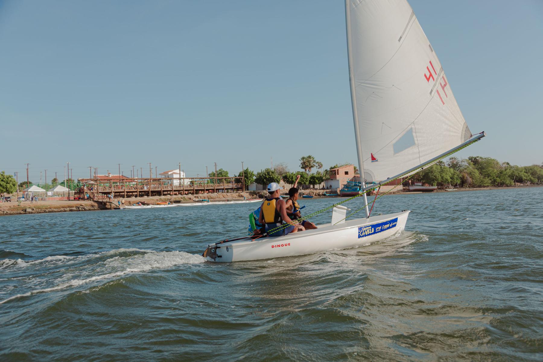 Dois jovens velejam um barco Dingue de vela branca no Rio Jaguaribe, com a orla de Aracati ao fundo